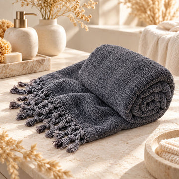 Antracietkleurige bamboe hamamdoek met franjes, opgerold op een natuurstenen blad in een lichte wellnessomgeving.