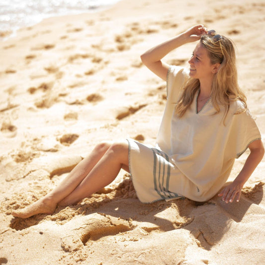 Vrouw zit ontspannen op het strand in een naturel katoenen strandponcho met korte mouwen en grijze strepen.