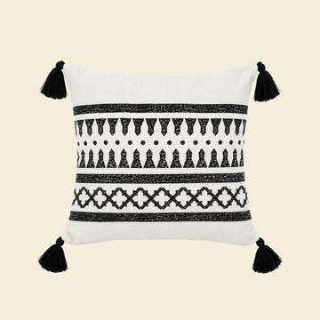 Witte kussenhoes met zwart etnisch patroon, geometrische motieven en zwarte tassels aan de vier hoeken met creme achtergrond.