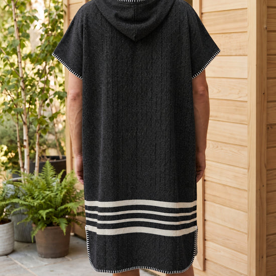 Zwarte sauna poncho met capuchon en witte strepen, gedragen door een man bij een houten sauna, achteraanzicht.