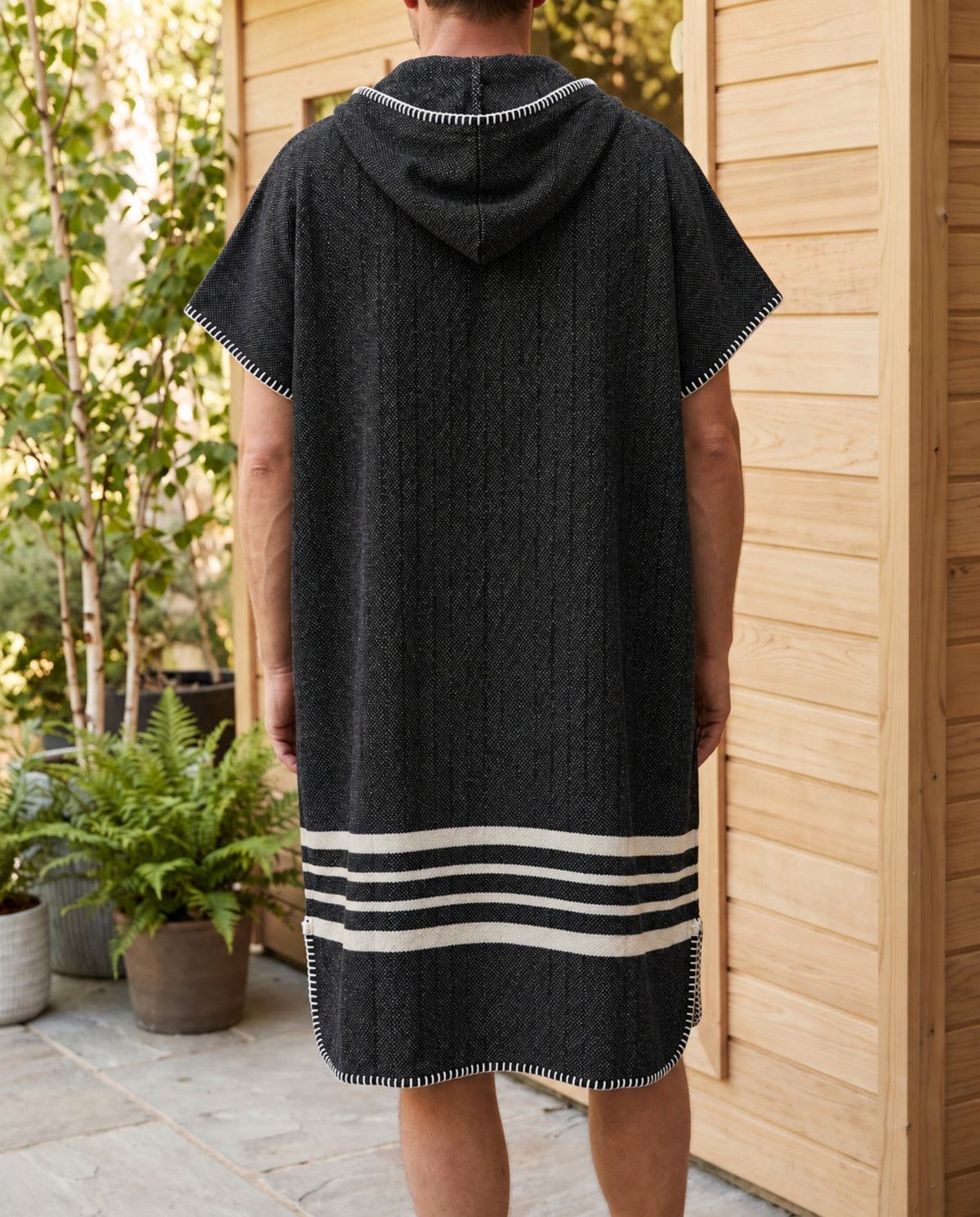Zwarte sauna poncho met capuchon en witte strepen, gedragen door een man bij een houten sauna, achteraanzicht.