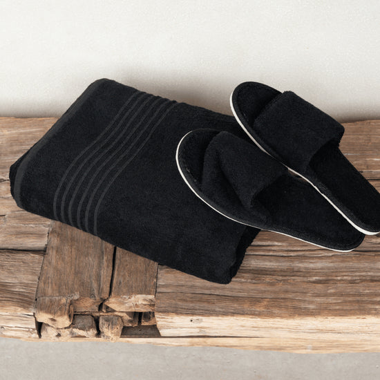 Zwart saunapakket met opgevouwen XXL saunadoek en zwarte saunaslippers op een rustieke houten bank.