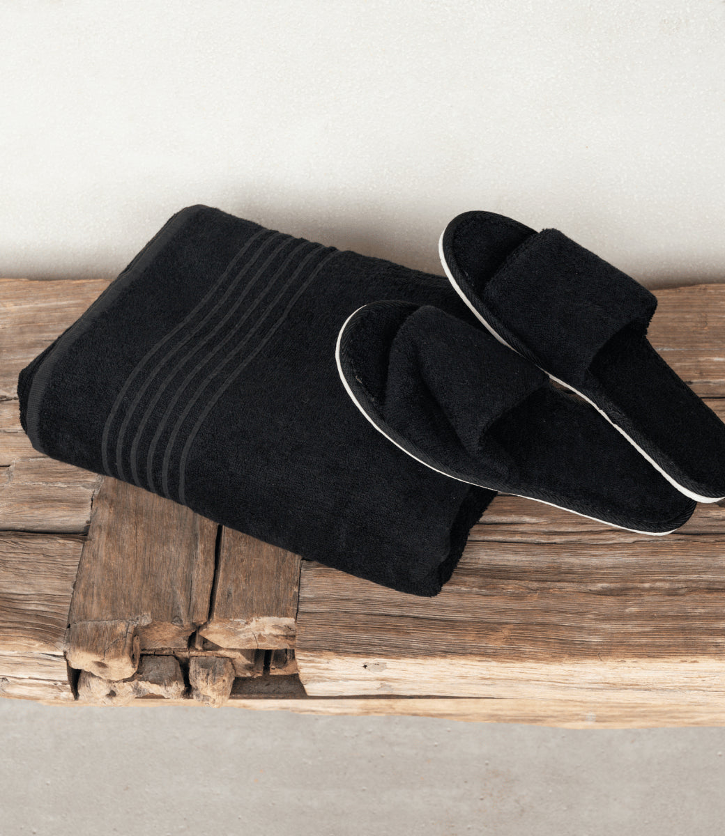 Zwart saunapakket met opgevouwen XXL saunadoek en zwarte saunaslippers op een rustieke houten bank.