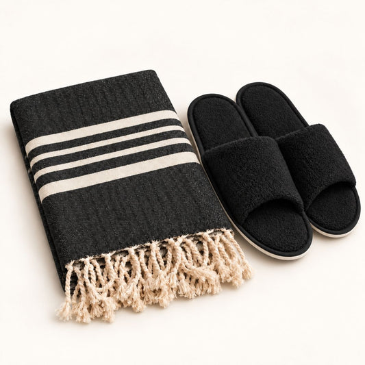 Saunaset met zwarte hamamdoek met naturelle strepen en franjes, gecombineerd met zwarte badstof saunaslippers met witte zoolrand.